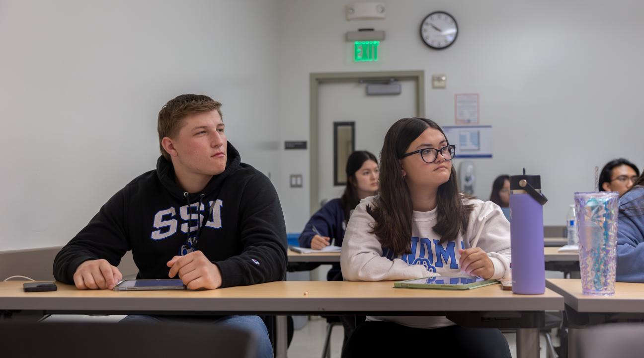 Two students in a classroom sitting and listening to someone not in view