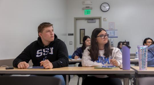 Two students in a classroom sitting and listening to someone not in view